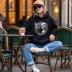 Puede incluir: Una sudadera negra con capucha que presenta un gr&aacute;fico circular de un Bulldog Franc&eacute;s y un perro tipo Doodle con el texto UGA & MIK impreso debajo. La sudadera tiene mangas largas, un bolsillo tipo canguro y se usa con pantalones vaqueros.
