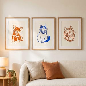Op de afbeelding: Drie ingelijste wandkunstprints met dierillustraties. De linker print toont een oranje Franse Bulldog, de middelste print een blauwe kat en de rechter print een oranje cyperse kat. Elk dier draagt een bandana met een patroon.