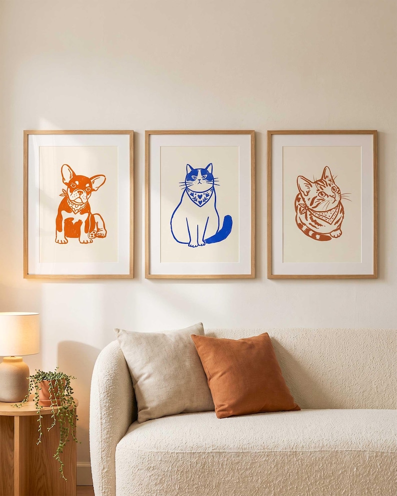 Op de afbeelding: Drie ingelijste wandkunstprints met dierillustraties. De linker print toont een oranje Franse Bulldog, de middelste print een blauwe kat en de rechter print een oranje cyperse kat. Elk dier draagt een bandana met een patroon.