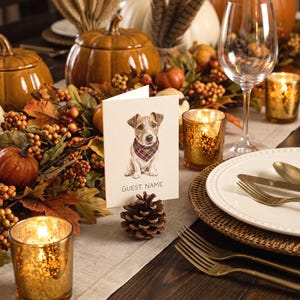 A folded white card featuring a watercolor illustration of a Jack Russell Terrier wearing a plaid bandana, with the text GUEST NAME printed below. The card is held upright by a natural brown pinecone on a table setting.