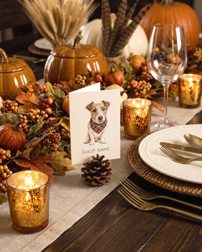 A folded white card featuring a watercolor illustration of a Jack Russell Terrier wearing a plaid bandana, with the text GUEST NAME printed below. The card is held upright by a natural brown pinecone on a table setting.