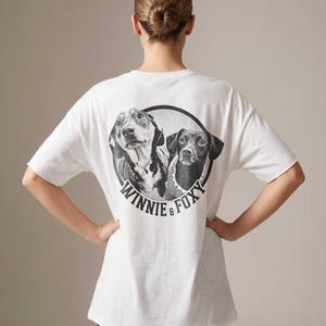 Puede incluir: Camiseta blanca con un dise&ntilde;o en blanco y negro de dos perros dentro de un c&iacute;rculo. El texto "WINNIE & FOXY" est&aacute; impreso debajo. La camiseta tiene mangas cortas y un corte holgado.