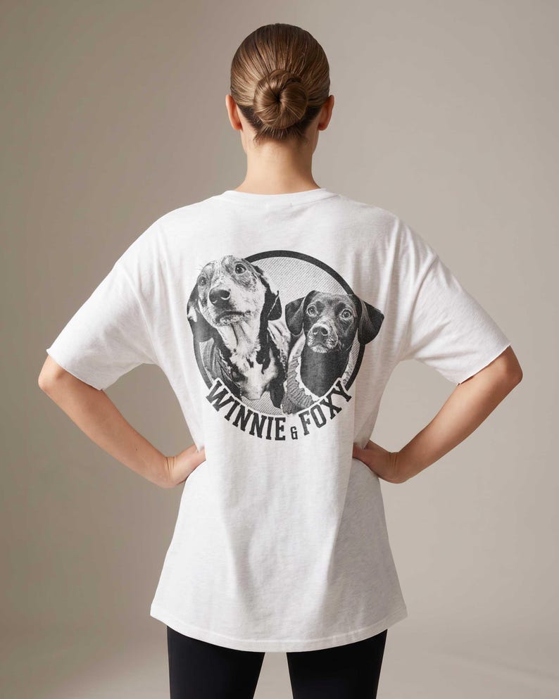 Puede incluir: Camiseta blanca con un dise&ntilde;o en blanco y negro de dos perros dentro de un c&iacute;rculo. El texto "WINNIE & FOXY" est&aacute; impreso debajo. La camiseta tiene mangas cortas y un corte holgado.