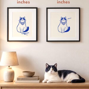 May include: Two framed wall art prints featuring a blue line drawing of a seated cat wearing a patterned bandana with hearts and leaves on a cream background. The left print is labeled 8x10 inches and the right print is labeled 10x10 inches.