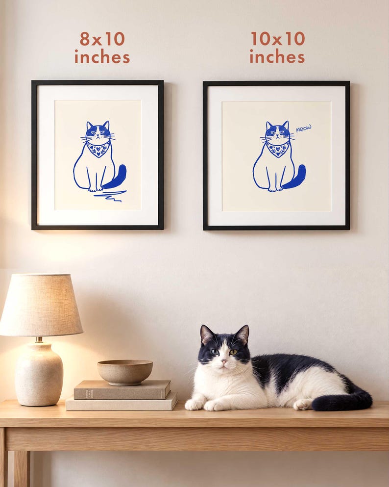 May include: Two framed wall art prints featuring a blue line drawing of a seated cat wearing a patterned bandana with hearts and leaves on a cream background. The left print is labeled 8x10 inches and the right print is labeled 10x10 inches.