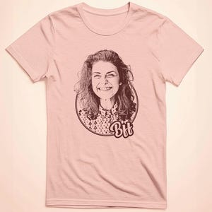 May include: A pink t-shirt featuring a black and white illustration of a woman smiling. The illustration is within a circle with the word "Bff" underneath.