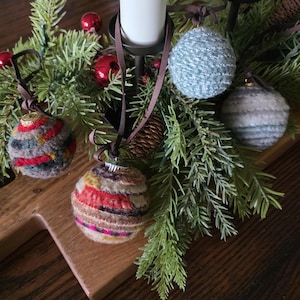 May include: Close-up of a holiday centerpiece featuring four decorative, yarn-wrapped ornaments in various colors. The ornaments hang from brown ribbons and are interspersed with evergreen branches, red berries, and a candle holder.