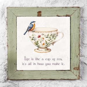 May include: A white teacup with floral designs and a blue bird perched on the rim. The teacup is on a white background with the text "Life is like a cup of tea, it's all in how you make it."
