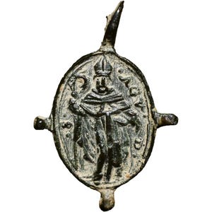 Op de afbeelding: Antieke ovale bronzen hanger met een gedetailleerde afbeelding van een geklede figuur met een mijter. De hanger heeft een lus aan de bovenkant en twee kleine uitsteeksels aan de zijkanten. De figuur is omringd door tekst.
