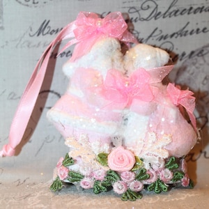 May include: A decorative ornament featuring a pair of pink and white baby booties. The ornament is adorned with pink bows, lace, and a floral base with green leaves and pink roses. A pink ribbon is attached for hanging.