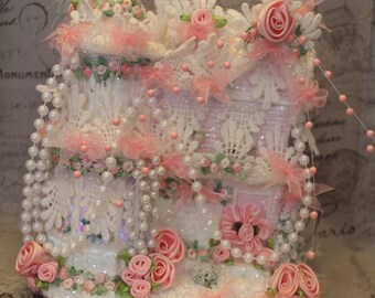 Puede incluir: Una casa en miniatura blanca y rosa decorada con encaje, perlas y flores rosas. La casa está cubierta de purpurina y tiene una base nevada.