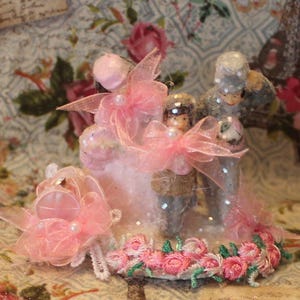 May include: A miniature wedding cake topper featuring a bride and groom figurine. The cake is decorated with pink bows, pearls, and a garland of pink embroidered roses. The figures are dressed in gray and brown attire.