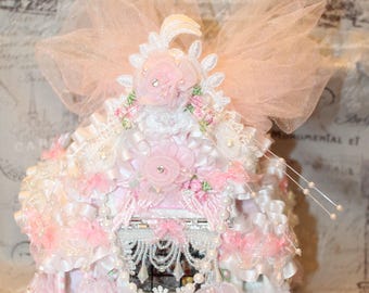 Puede incluir: Una caja decorativa y ornamentada en tonos rosa y blanco, adornada con encaje, cintas y adornos florales. La caja presenta un delicado diseño de cortina y está rematada con un gran lazo rosa transparente. Perlas y pedrería añaden brillo.