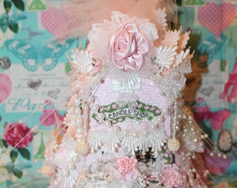 Puede incluir: Una casa rosa en miniatura decorativa con el texto "CANDLE SHOP" en la parte delantera. La casa está adornada con encaje, perlas y flores de tela rosa. Una corona de tul rosa y rosas de satén corona la parte superior de la casa.