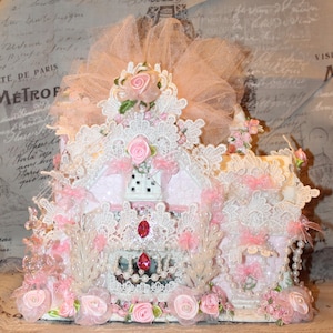 May include: A pink and white miniature house decorated with lace, tulle, and pink flowers. The house has a glittery white exterior and a pink roof. The house is surrounded by pink flowers and greenery.