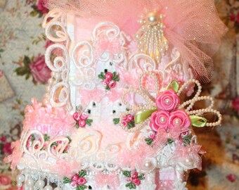Puede incluir: Una casa en miniatura decorativa, predominantemente rosa y blanca, adornada con encaje, perlas y detalles florales. El techo está rematado con tul rosa y un adorno de perlas. La casa presenta múltiples niveles y detalles intrincados.