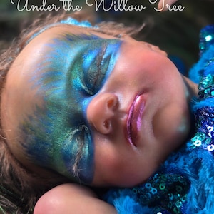 Może przedstawiać: Śpiąca lalka niemowlęca z niebieskim i zielonym makijażem oczu i niebieskim, cekinowym kostiumem. Lalka leży na łóżku z zielonych i niebieskich piór. Tekst "Under the Willow Tree" jest napisany nad lalką.