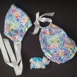 May include: A baby outfit set with a white onesie, a white hat, and a white headband with a blue bow. The onesie and hat have a colorful swirl pattern.