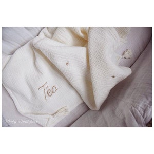 May include: A white muslin baby blanket with the name "Tea" embroidered in brown thread. The blanket has tassels on the corners.