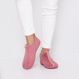 May include: Pink suede slippers with cream-coloured lining and trim. The slippers have a low-cut design and are worn with white trousers. The slippers appear soft and comfortable, suitable for indoor wear.