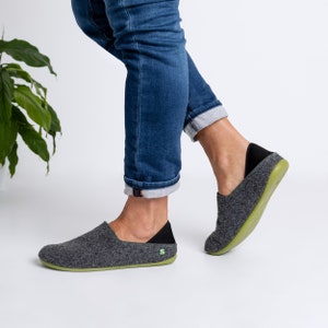 May include: Grey felt slip-on shoes with a black heel counter and a green sole. The shoes have a minimalist design and a small green logo on the side. The shoes are shown with blue jeans.