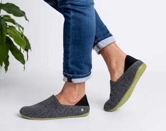 May include: Grey felt slip-on shoes with a black heel counter and a green sole. The shoes have a minimalist design and a small green logo on the side. The shoes are shown with blue jeans.