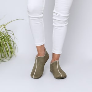 Könnte beinhalten: Ein Paar olivgrüne Slipper aus Wildleder mit weißem Besatz. Die Slipper haben eine abgerundete Spitze und eine flache Sohle.