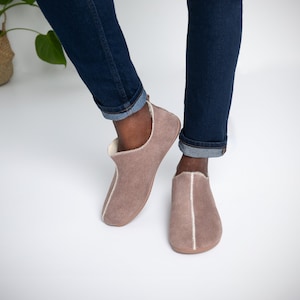 May include: A pair of light brown suede slippers with a white trim. The slippers are worn by a person wearing blue jeans.