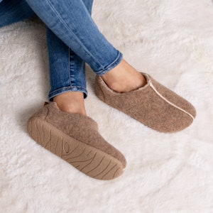 May include: A pair of brown slippers with a light-colored trim and sole. The slippers are made of a soft, textured material and have a rounded toe. The soles are a darker brown with a textured pattern. The slippers are worn with blue jeans.