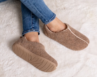 May include: A pair of brown slippers with a light-coloured trim and sole. The slippers are made of a soft, textured material and have a rounded toe. The soles are a darker brown with a textured pattern. The slippers are worn with blue jeans.