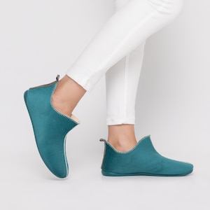 May include: Teal suede ankle boots with a cream interior and stitched trim. The low-profile boots have a small pull tab at the heel. The boots are shown on a person wearing white trousers.