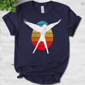 Puede incluir: Una camiseta azul marino con una silueta blanca de un pájaro volando frente a un diseño de puesta de sol retro.