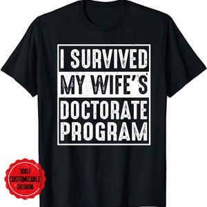 Könnte beinhalten: Schwarzes T-Shirt mit weißem Text, der "Ich habe das Doktorat meiner Frau überlebt" lautet.