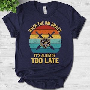 May include: A navy blue t-shirt featuring a vintage-style graphic of a 20-sided die and crossed swords. The text "When the DM smiles it's already too late" is printed on the shirt.