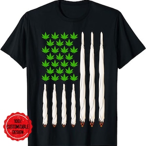 Op de afbeelding: Een zwart T-shirt met een groen marihuanabladontwerp in de vorm van de Amerikaanse vlag. De bladeren zijn in rijen gerangschikt en de witte strepen van de vlag worden weergegeven door witte, opgerolde marihuanajoints.