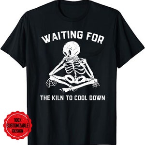 Op de afbeelding: Zwart T-shirt met een wit skelet dat met zijn kin op zijn hand zit. De tekst "WAITING FOR THE KILN TO COOL DOWN" staat op het T-shirt gedrukt.