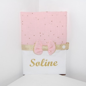 Peut inclure: Un livre de bébé rose et or avec un nœud et le nom "Soline" écrit en paillettes dorées.
