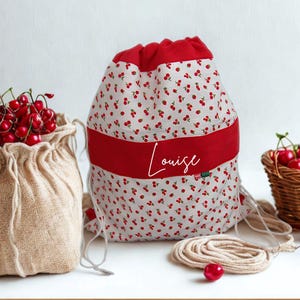 Peut inclure: Un sac à cordon avec une bande rouge et un imprimé cerise, avec le nom "Louise" en écriture blanche. Un sac en toile de jute et un panier en osier, tous deux remplis de cerises rouges fraîches, sont également dans l'image.