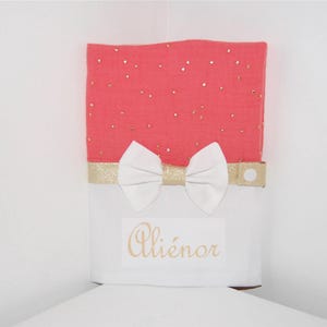 Peut inclure: Une bavette en mousseline rose corail avec des points de paillettes dorés et un nœud en satin blanc. La bavette est ornée d'une bordure dorée et d'une fermeture à pression. La bavette est personnalisée avec le nom "Aliénor" en broderie dorée.