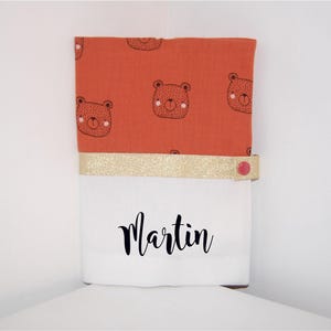 Peut inclure: Une pochette en tissu blanc avec une bordure dorée et un bouton rouge. La pochette a un dessus en tissu rouge avec un motif d'ours bruns. La pochette porte le nom "Martin" sur le devant.