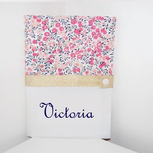 Peut inclure: Une boîte blanche avec un motif floral sur le dessus avec des fleurs roses et des feuilles vertes. Une bande dorée à paillettes sépare le dessus floral de la base blanche. Le nom "Victoria" est écrit en script bleu marine.