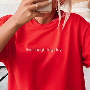Puede incluir: Camiseta roja de manga corta con el texto "live, laugh, leg day" impreso en blanco en la parte delantera.