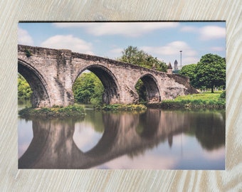 May include: A stone bridge with three arches spans a river, reflecting the structure in the still water. Lush green trees line the riverbanks, and a tall tower stands in the distance.