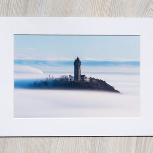 May include: A stone tower stands on a hilltop, surrounded by a sea of fog. The sky is a soft blue, with wispy clouds.