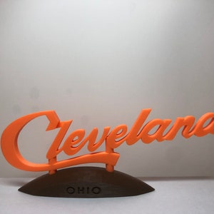 May include: Orange 3D printed sign that reads "Cleveland, Ohio" in a cursive font. The sign is mounted on a dark brown base.