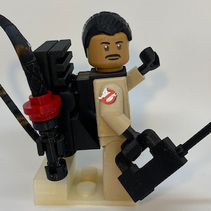 May include: A tan and black Lego-style figure of a Ghostbuster, complete with a proton pack and a black weapon. The figure has a mustache and the Ghostbusters logo on the chest. The figure is standing on a light tan base.