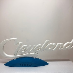 May include: White 3D printed sign that reads "Cleveland Ohio" in a cursive font. The sign is mounted on a blue base.