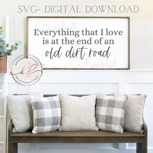 May include: A white sign with black text that reads "Everything that I love is at the end of an old dirt road" with the words "Barefoot & Freckled" written in cursive at the top and bottom of the sign. The sign is hanging on a white wall above a wooden bench with gray and white striped pillows.