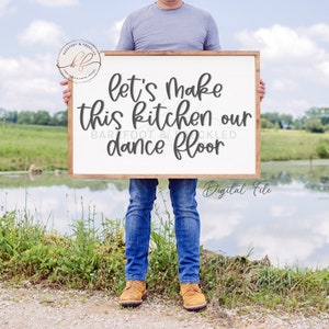 May include: A framed wooden sign with the text "Let's make this kitchen our dance floor" in a handwritten style. The sign is held by a person in blue jeans and work boots, with a lake and greenery in the background.
