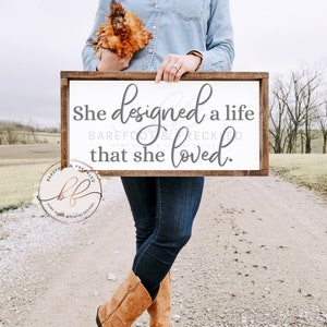 May include: A white wooden sign with the text "She designed a life that she loved."  The sign is held by a person wearing a blue denim shirt and brown cowboy boots. The text "BAREFOOT & FRECKLED" is also on the sign.